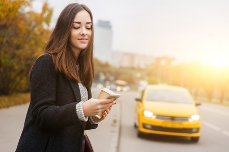 Beautiful brunette with phone in hand standing in street on background of passingyellow taxiの写真素材