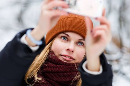 Blonde girl in scarf and hat making selfie on street in winterの写真素材