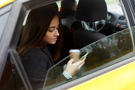 Young woman with coffee and phone in hand coming out of taxiの写真素材