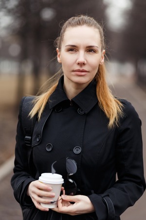Beautiful girl in a black raincoat and with coffee in handsの写真素材