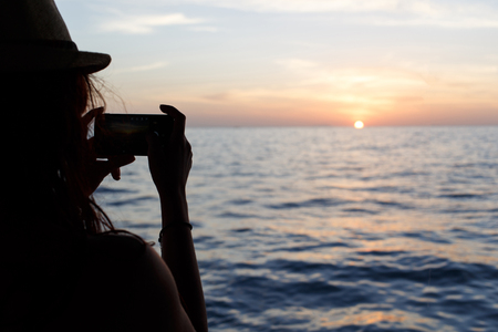 Girl in hat taking pictures of sunset over seaの写真素材
