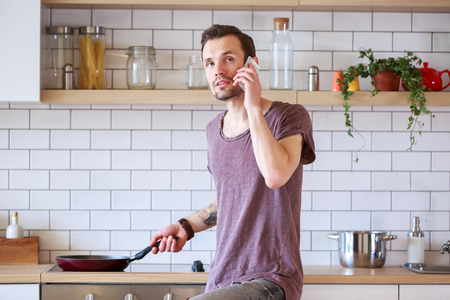 Photo of brunet man with frying pan in his hands talking on phone in kitchenの写真素材