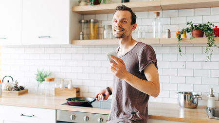 Portrait of brunet man with frying pan in his hands talking on phone in kitchenの写真素材