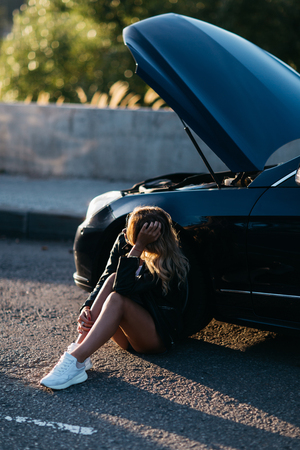 Photo of upset woman sitting on asphalt next to broken car with open hoodの写真素材