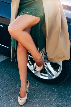 Photo of legs of woman in dress and raincoat close-up standing by carの写真素材