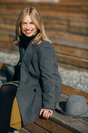 Portrait of young blonde in gray coat sitting on wooden benchの写真素材