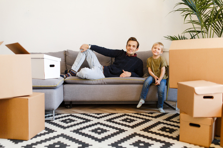Photo of happy father and son sitting on gray sofa among cardboard boxesの写真素材