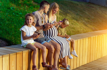 Image of parents and two children sitting on wooden fence with phones in hands in park