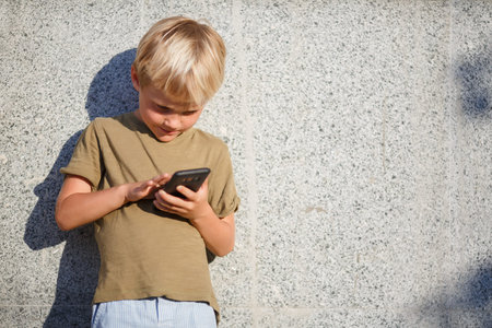 Photo of boy with phone in his hands standing near concrete wall