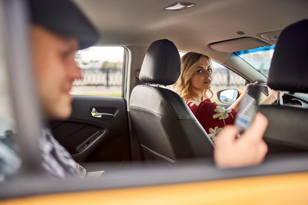 Photo of young man in cap with phone in hand sitting in back seat, women driving in yellow taxi