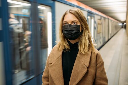 Blonde woman in black medical mask looking at camera while standing next to subway car.の写真素材