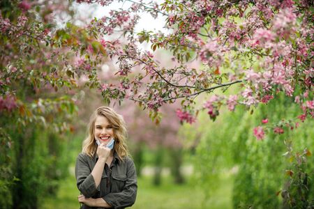 Young woman in medical mask on background of flowering trees in park at afternoon.の写真素材