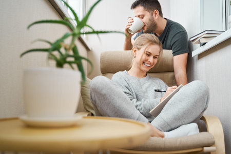 Happy blonde woman sitting on beige armchair and brunet man standing beside her in apartment