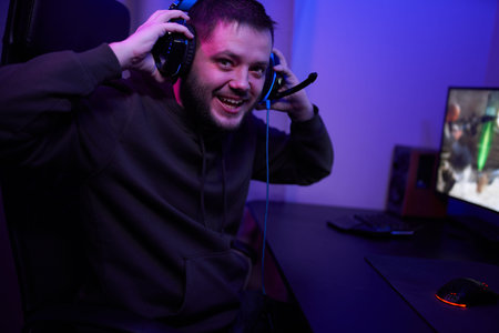 Gamer or streamer in headphones with microphone sits at home in neon light and plays at pc