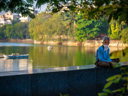 Thane, India, December 22,2020 :old aged Indian man sitting beside a lake covering his face with aÂ handkerchiefÂ to protect from Covid-19のeditorial素材
