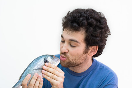 Young man kissing a fish. Curly hair with beard. White caucasian. Isolated on white.