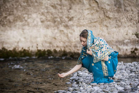 The girl in the Indian sari sits at the small river and drinks from handsの写真素材