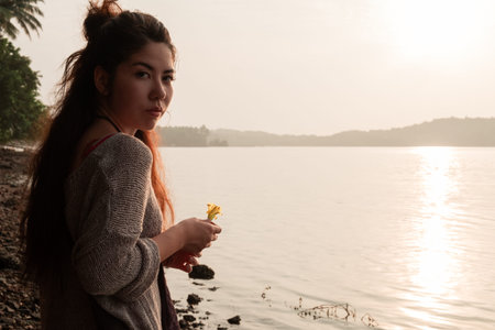 Asian girl stands on the seashore with a flower in her hands, meets the sunset