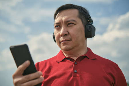 Middle-aged Asian man in headphones outdoors listening to music against the background of the sky, mobile phone in his handの写真素材