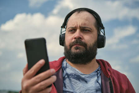 Middle-aged European man in headphones outdoors listening to music against the background of the sky, mobile phone in his handの写真素材