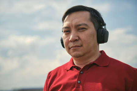 Middle-aged Asian man in headphones outdoors listening to music against the background of the skyの写真素材