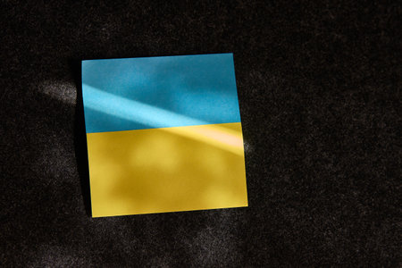 The sticker in the form of the Ukrainian flag lies. on a dark surface and a ray of sunlight falls on itの写真素材