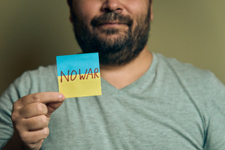 A bearded European man holds a sticker in front of him, in the form of a Ukrainian flag with the inscription no warの写真素材