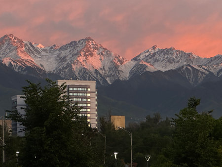 Scenic view of snow-covered mountains glowing in the sunset behind a cityscape with lush greenery. Perfect blend of nature and urban life.の写真素材