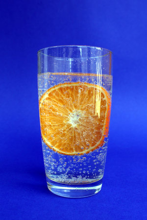 There is a transparent glass with water and an orange on a blue background