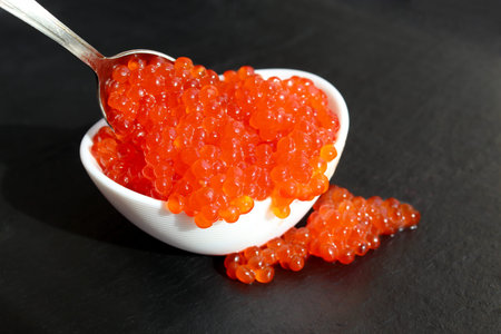 Red caviar in a white plate with a spoon lies on a black background