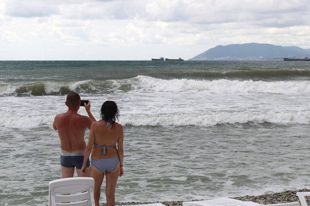 A guy and a girl stand and watch the raging sea. The guy takes pictures of the waves on the phone.の写真素材
