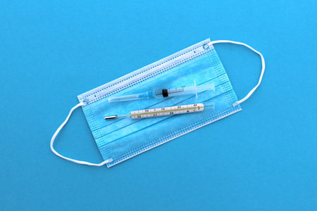 On a medical mask lie a thermometer and a syringe with medicine on a blue background.
