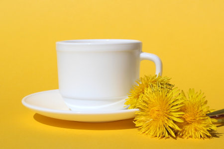 On a yellow background stands a white cup with a saucer and dandelion flowers.の写真素材