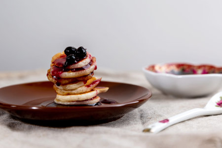 A stack of mini pancakes on a brown saucer with blackcurrant jam.