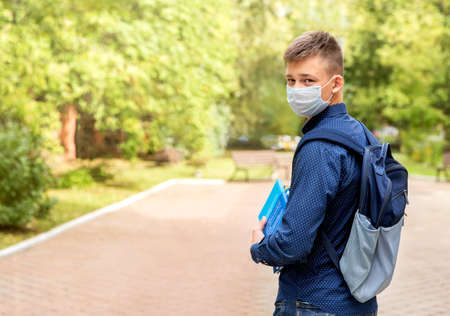 A teenager in a medical mask with books in his hands in the schoolyard. He turned around. He has school books in his hands and a backpack on his back.の写真素材