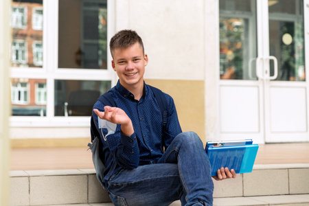One teenage schoolboy is sitting on the steps of his school. Hes holding a medical mask. He smiles....