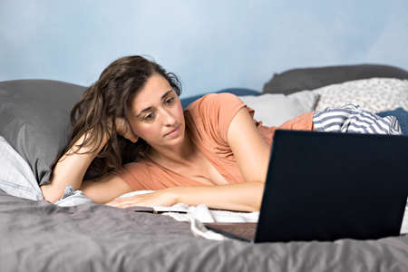 A beautiful brunette woman in her bedroom is lying on the bed and looking at her laptop. She's resting. The girl is watching a movie or video tutorial.の写真素材