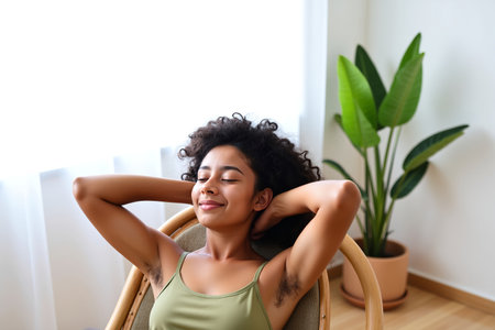 A young woman with her arms behind her head smiles while sitting in a chair near a windowの素材