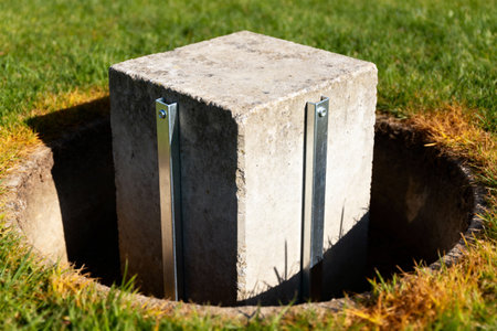 A concrete cube is secured in a circular hole in the ground with metal supports, surrounded by green grassの素材