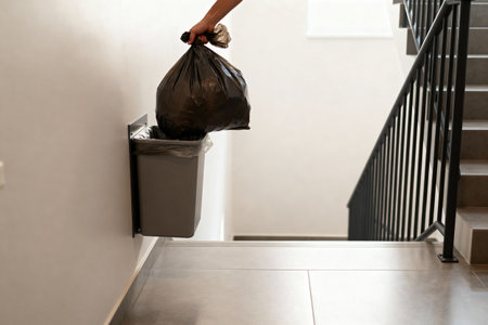 A hand is holding a black trash bag above a waste bin, ready to dispose of the garbageの写真素材