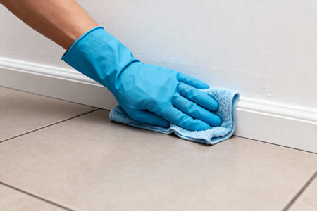 A person wearing a blue glove cleans a white baseboard with a blue cloth, promoting cleanliness and...
