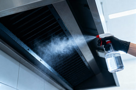 A glove hand sprays a cleaning solution onto a commercial kitchen exhaust hood, promoting hygiene and cleanlinessの写真素材