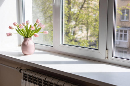 A delicate bouquet of pale pink tulips sits in a textured pink vase on a bright white windowsill, with dappled sunlight creating shadows and a blurred view of green trees and buildings outsideの写真素材