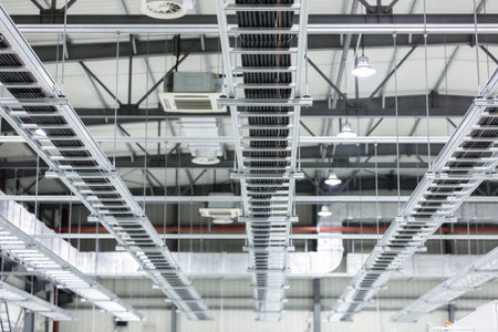 Rows of metal cable trays carrying wires are suspended from the ceiling of a modern industrial...