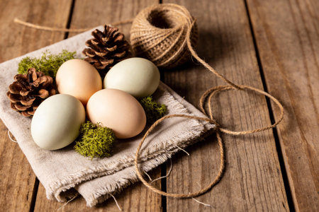 A rustic arrangement of pastel-colored Easter eggs, pine cones, and green moss on a textured linen...