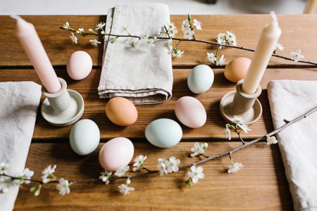 A rustic wooden table is adorned with pastel-colored Easter eggs, delicate blooming branches, and minimalist candles, creating a serene spring atmosphereの写真素材