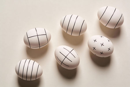 Six white eggs adorned with geometric black line designs are artfully arranged on a soft, neutral...