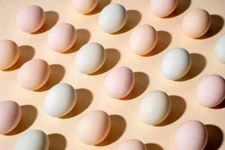 A collection of pastel colored eggs laid out in a symmetrical grid formation on a light peach background, casting subtle shadowsの写真素材