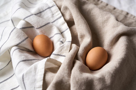A close-up view of two organic brown eggs resting on draped linen textiles, one with thin gray...