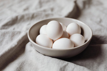 A collection of white eggs is presented in a simple ceramic bowl, set against a backdrop of soft,...